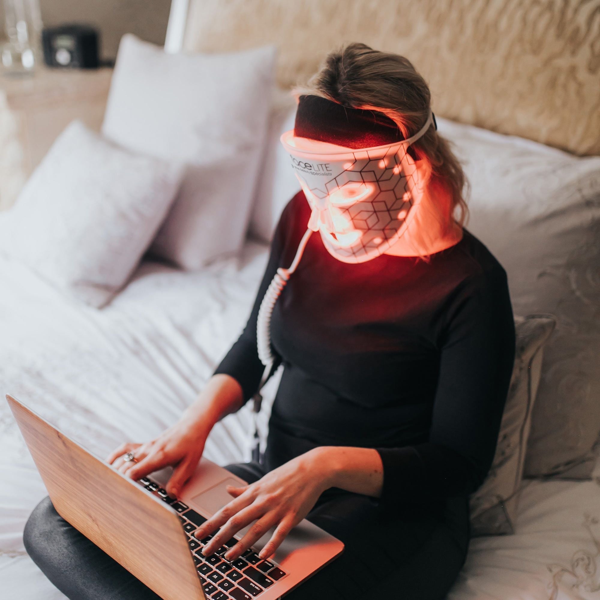 lady dressed in black relaxing at home on bed using laptop wearing facelite beauty boosting LED face mask with red and infrared lights illuminated 