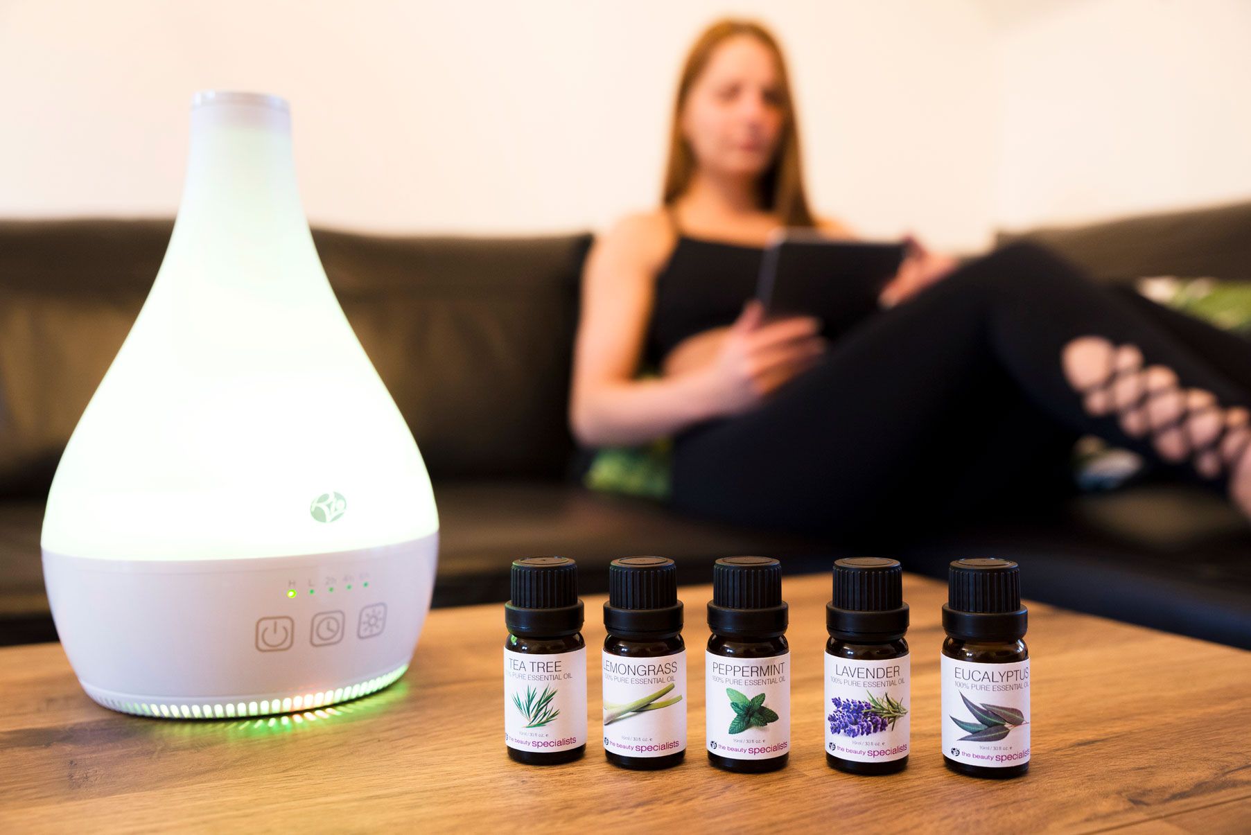 rio aromatherapy 100% essential oil collection placed on table next to diffuser in a home setting with lady relaxing on sofa in the background
