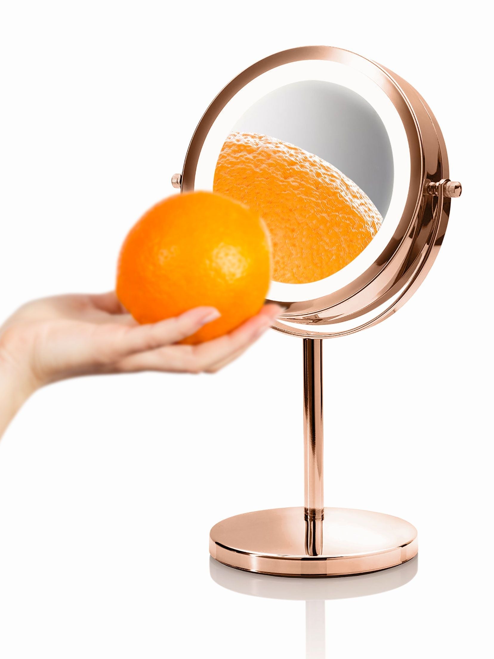 rose gold double sided cosmetic mirror with orange held in front of it with magnified reflection of orange peel